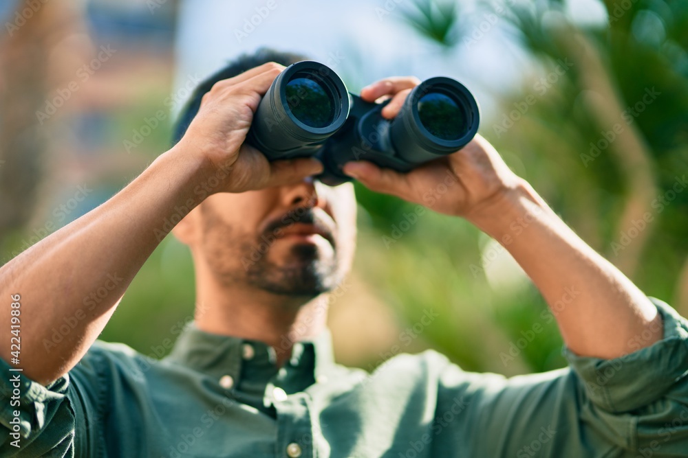 Young hispanic man smiling happy looking for new opportunity using binoculars at the park. Young hispanic man smiling happy looking for new opportunity using binoculars at the park.
