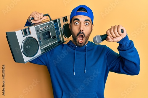Young hispanic man holding boombox, listening to music singing with microphone afraid and shocked with surprise and amazed expression, fear and excited face.