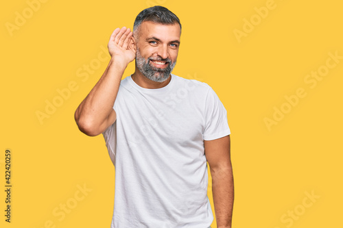 Middle age handsome man wearing casual white tshirt smiling with hand over ear listening an hearing to rumor or gossip. deafness concept.