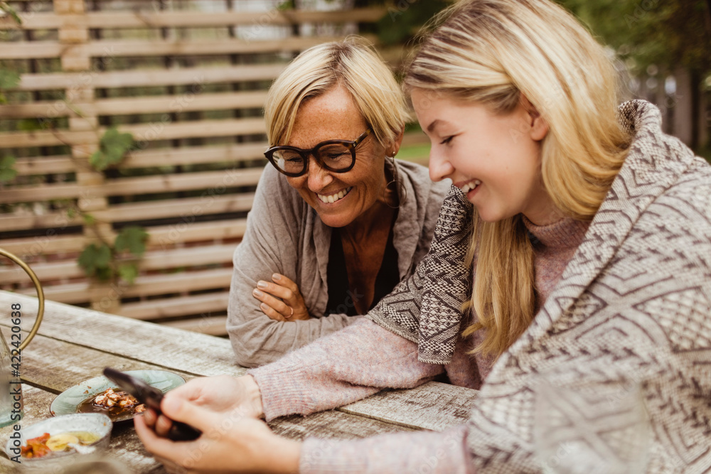 Smiling granddaughter using smart phone sitting with grandmother in front yard