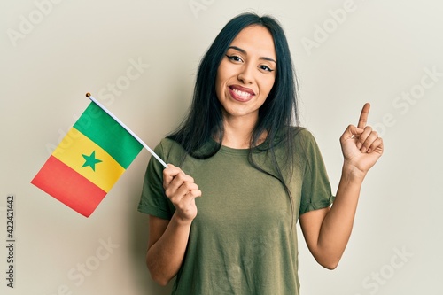 Young hispanic girl holding senegal flag smiling happy pointing with hand and finger to the side