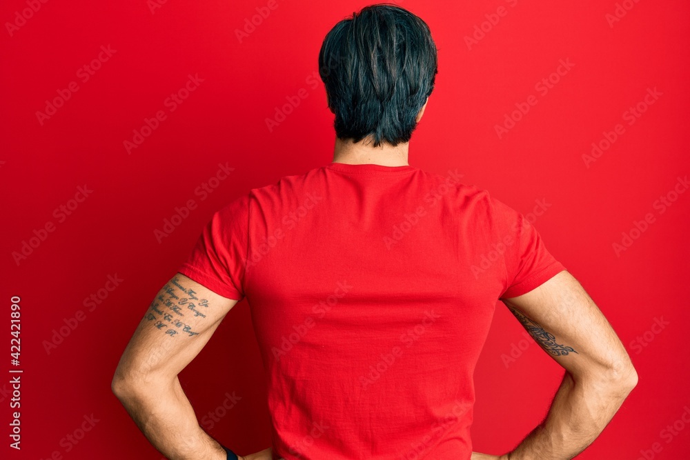Young hispanic man wearing casual red t shirt standing backwards looking away with arms on body Young hispanic man wearing casual red t shirt standing backwards looking away with arms on body