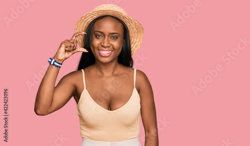 Young black woman wearing summer hat smiling and confident gesturing with hand doing small size sign with fingers looking and the camera. measure concept. © Krakenimages.com