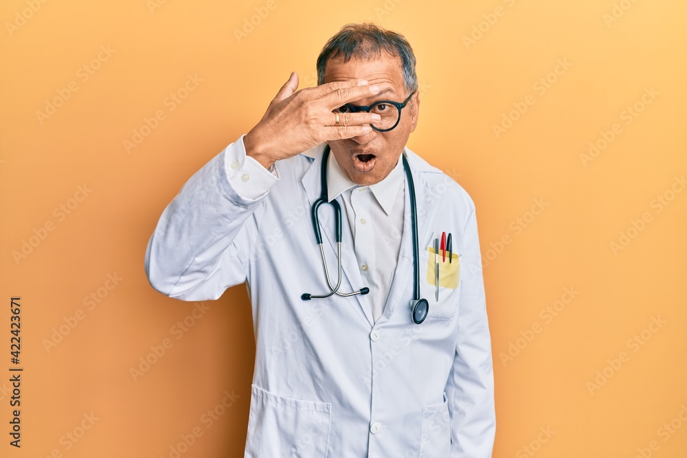 Middle age indian man wearing doctor coat and stethoscope peeking in shock covering face and eyes with hand, looking through fingers with embarrassed expression. Middle age indian man wearing doctor coat and stethoscope peeking in shock covering face and eyes with hand, looking through fingers with embarrassed expression.