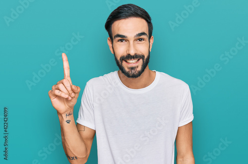 Young hispanic man wearing casual white t shirt showing and pointing up with finger number one while smiling confident and happy.