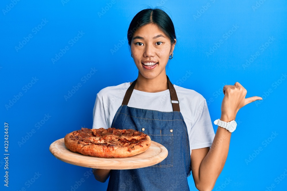 Young chinese woman wearing waiter apron holding italian pizza pointing thumb up to the side smiling happy with open mouth Young chinese woman wearing waiter apron holding italian pizza pointing thumb up to the side smiling happy with open mouth