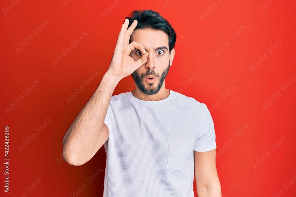 Young hispanic man wearing casual white tshirt doing ok gesture shocked with surprised face, eye looking through fingers. unbelieving expression. Young hispanic man wearing casual white tshirt doing ok gesture shocked with surprised face, eye looking through fingers. unbelieving expression.