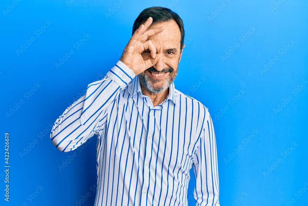 Middle age hispanic man wearing casual clothes doing ok gesture with hand smiling, eye looking through fingers with happy face. Middle age hispanic man wearing casual clothes doing ok gesture with hand smiling, eye looking through fingers with happy face.