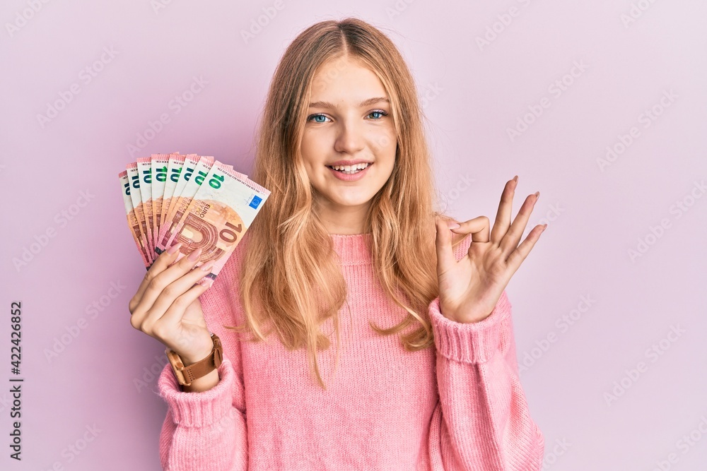 Obraz premium Beautiful young caucasian girl holding bunch of 10 euro banknotes doing ok sign with fingers, smiling friendly gesturing excellent symbol