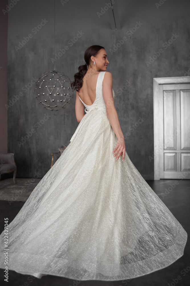 Pretty young caucasian brunette bride with make-up and pony tail hairstyle in long wedding ball gown dress standing and posing in grey interior Pretty young caucasian brunette bride with make-up and pony tail hairstyle in long wedding ball gown dress standing and posing in grey interior