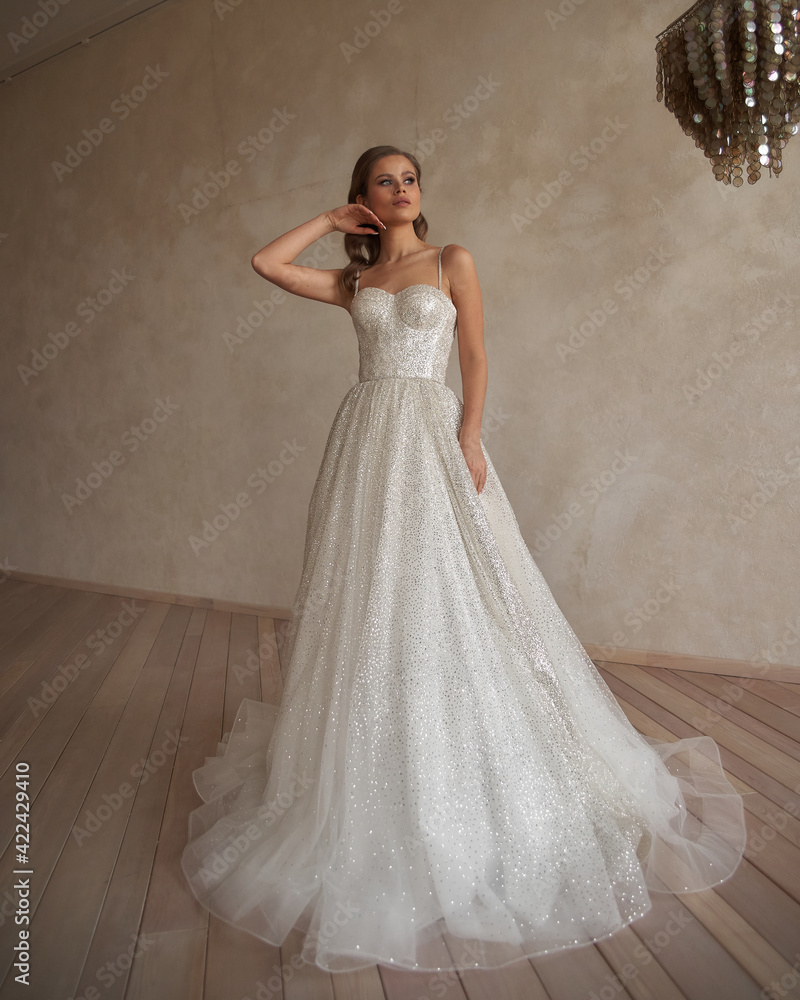 Fototapeta premium Full length portrait of young beautiful woman wearing white wedding dress. Elegant bride standing and posing against beige wall