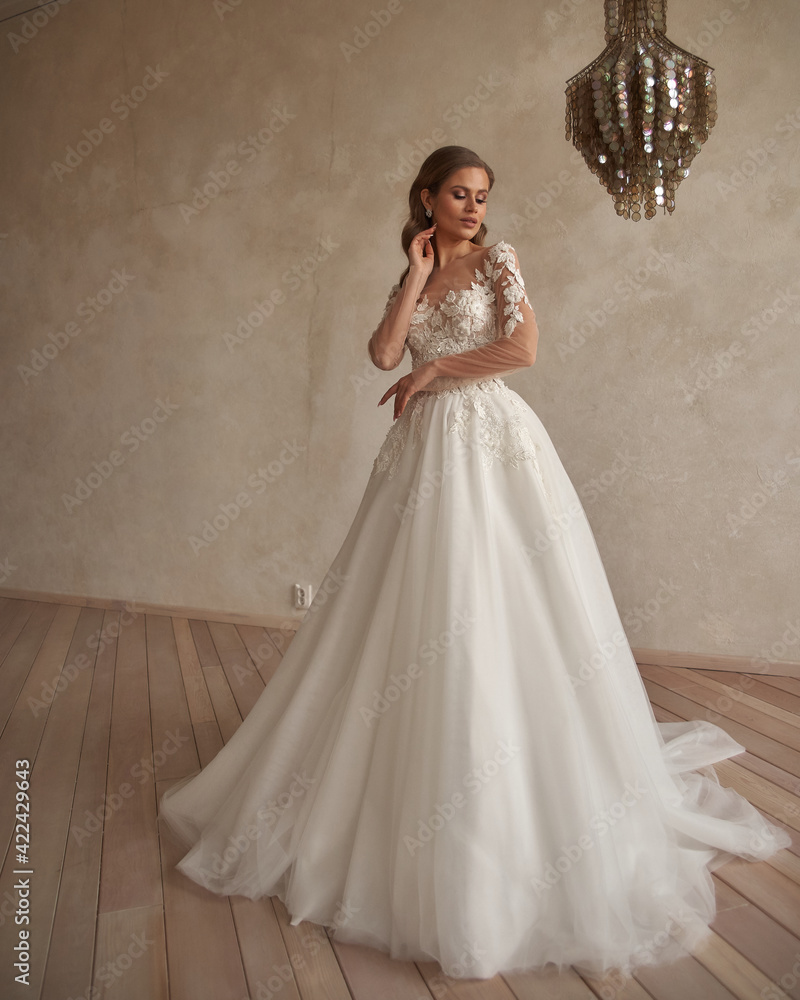 Full length portrait of young beautiful woman wearing white wedding dress. Elegant bride standing and posing against beige wall Full length portrait of young beautiful woman wearing white wedding dress. Elegant bride standing and posing against beige wall