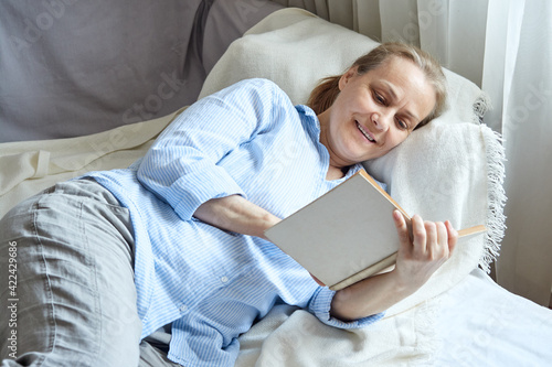 Smiling senior woman reading book while lying on sofa at home at free time.