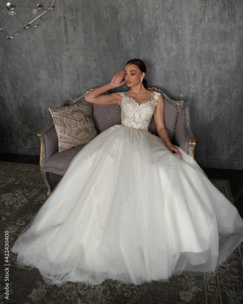 Pretty bride in white wedding dress. Young woman with perfect natural make-up and pony tail hairstyle sitting at sofa in grey interior. Beautiful woman Pretty bride in white wedding dress. Young woman with perfect natural make-up and pony tail hairstyle sitting at sofa in grey interior. Beautiful woman