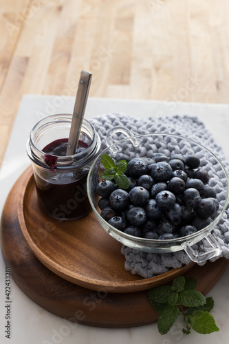A bowl full of fresh blueberries and a marmalade jar on a wooden tray with some mint leaves around.