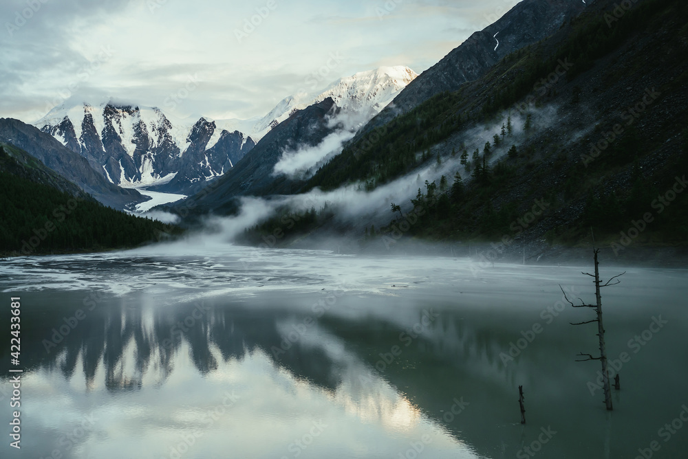 Scenic alpine landscape with snowy mountains in golden sunlight reflected on mirror mountain lake in fog among low clouds. Atmospheric highland scenery with low clouds on rocks and green mirror lake.