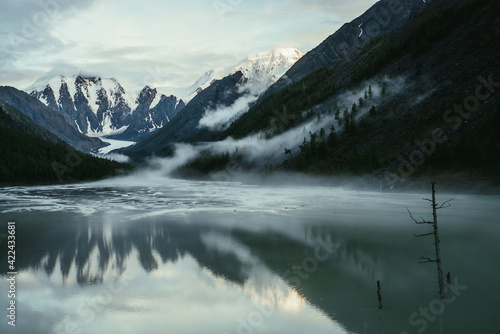 Scenic alpine landscape with snowy mountains in golden sunlight reflected on mirror mountain lake in fog among low clouds. Atmospheric highland scenery with low clouds on rocks and green mirror lake.