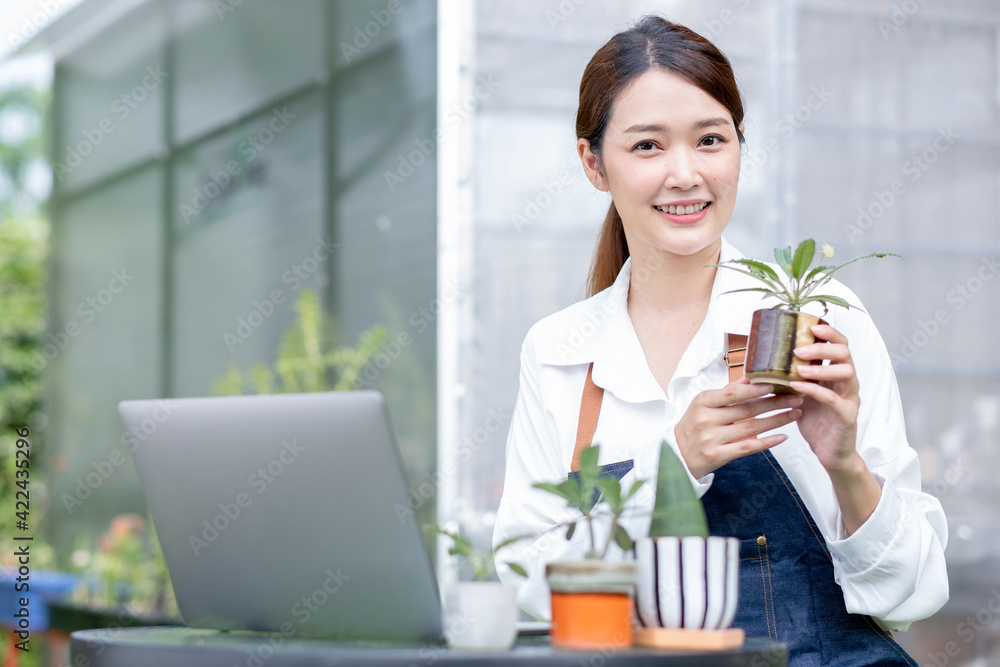 Beautiful Asian girl hold pot of plant and present to camera sit with laptop on table in front green garden of her house. Sustainable with grow cactus and other plant concept. Beautiful Asian girl hold pot of plant and present to camera sit with laptop on table in front green garden of her house. Sustainable with grow cactus and other plant concept.