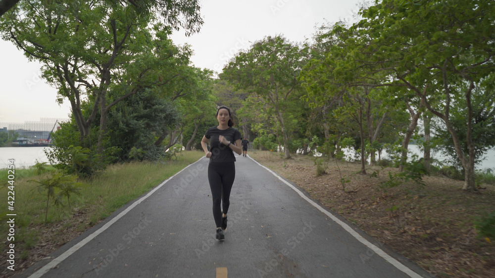 asian athlete woman in black sportswear and wireless headphone running in urban city trees park during sunset and bad cloudy weather. female jogging outdoor in evening. healthy workout.