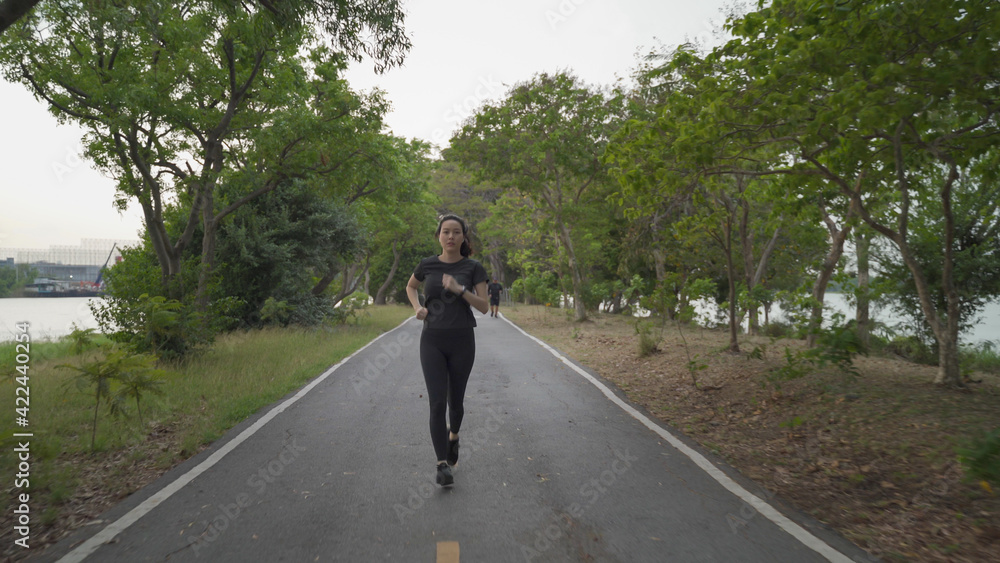 asian athlete woman in black sportswear and wireless headphone running in urban city trees park during sunset and bad cloudy weather. female jogging outdoor in evening. healthy workout. asian athlete woman in black sportswear and wireless headphone running in urban city trees park during sunset and bad cloudy weather. female jogging outdoor in evening. healthy workout.