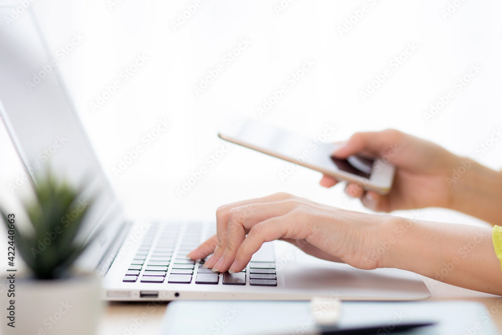 Fototapeta premium Closeup hand of young asian businesswoman working on laptop computer on desk at home office, freelance woman looking message on smart phone and typing on notebook, business and communication concept.