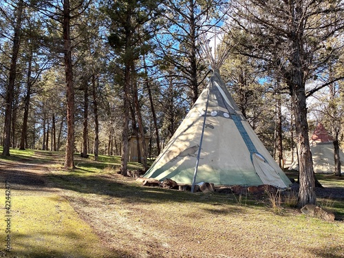 A teepee on a hill by a path through the trees in a campground on a sunny day.