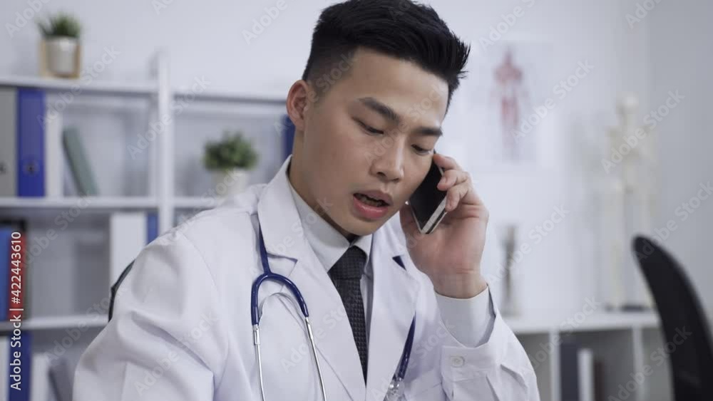 closeup handsome asian doctor wearing white coat is holding and talking on the smartphone at background contemporary medical workplace. closeup handsome asian doctor wearing white coat is holding and talking on the smartphone at background contemporary medical workplace.