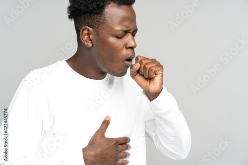 Sick African American man strongly coughing. Black man feeling unwell, suffered from asthma, flu, allergy, bronchitis, tuberculosis, virus, touching her chest, isolated on studio grey white background photo