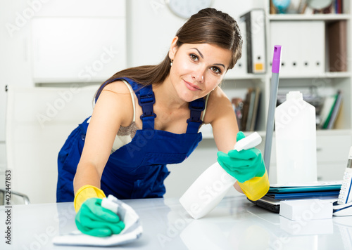 Young woman in an overalls cleans the table at office. High quality photo