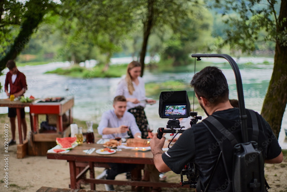 a professionally equipped cameraman is filming a group of people having dinner by the river. a professionally equipped cameraman is filming a group of people having dinner by the river.