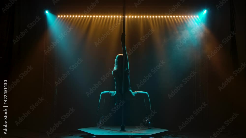 Sexy slim woman dancing striptease and spinning on a pole in a nightclub. Beautiful hot blonde stripper erotically moves on stage with neon lighting. Silhouette. Slow motion.