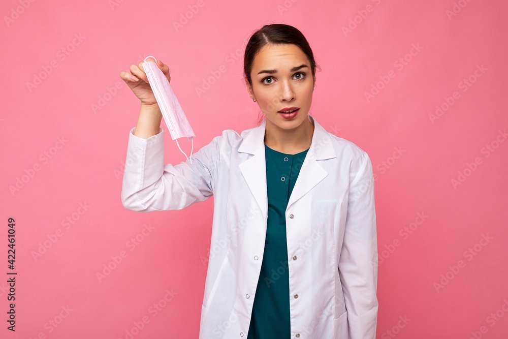 Portrait of a dissatisfied attractive young female doctor in white coat holding medical mask standing isolated on pink background Portrait of a dissatisfied attractive young female doctor in white coat holding medical mask standing isolated on pink background