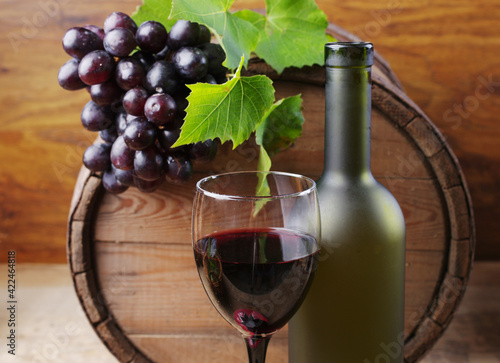 A glass of red wine, a bottle of wine against the background of a wine barrel, decorated with a branch of grapes.