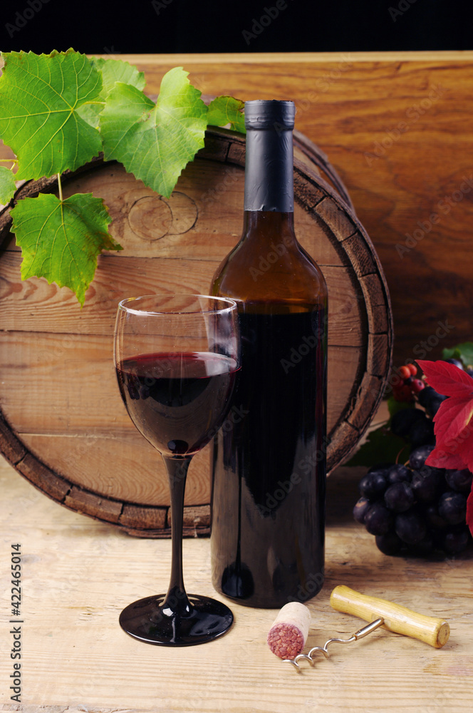 A glass of red wine, a bottle of wine against the background of a wine barrel, decorated with a branch of grapes.