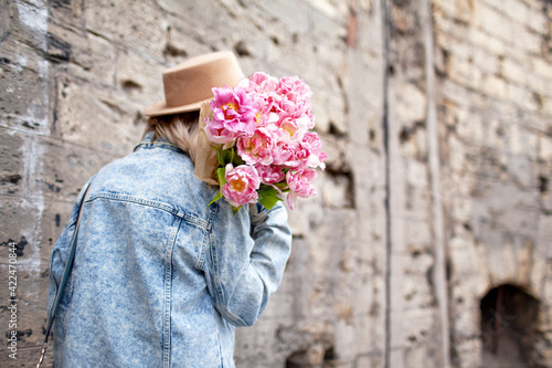 Fashion style woman dressed jeans jacket, hat and red dress walking in the street of old city with bouquet of flowers