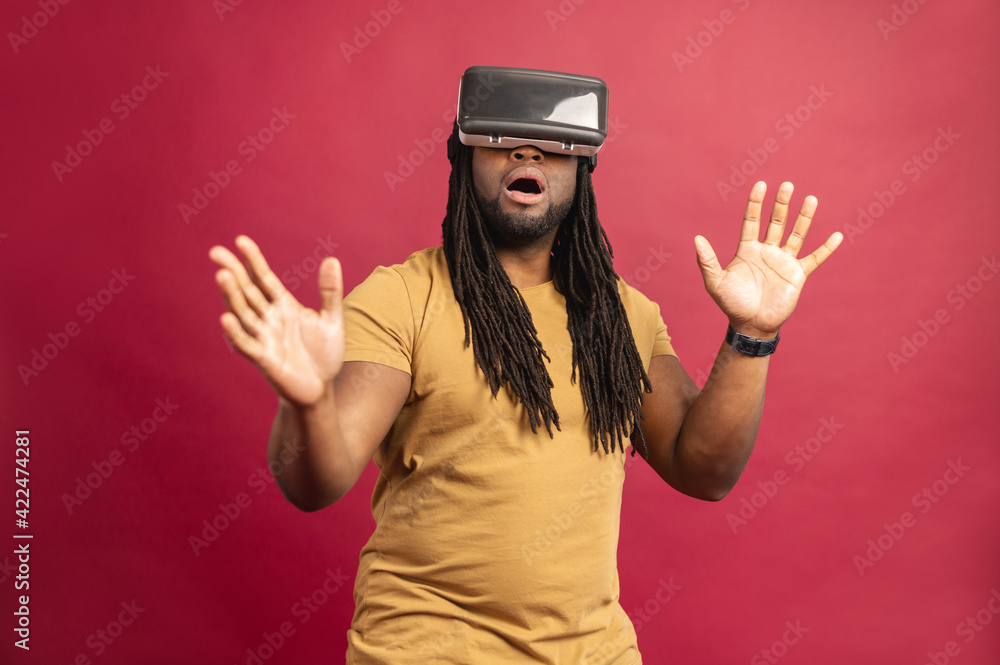 Surprised scared African American mixed-race man with dreadlocks and open mouth isolated over red wall touching air with hands, spending time in virtual reality, playing games in modern VR headset Surprised scared African American mixed-race man with dreadlocks and open mouth isolated over red wall touching air with hands, spending time in virtual reality, playing games in modern VR headset