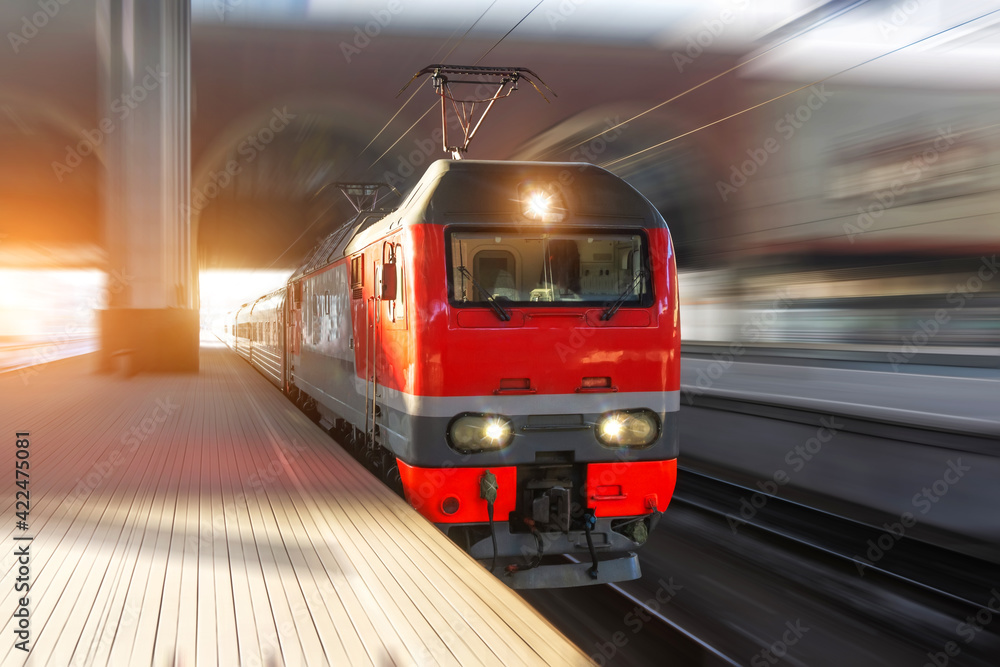 Fototapeta premium The lead electric locomotive with a passenger train passes at high speed through the city platform at the station.