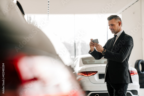 Mature man taking photo on cellphone while choosing car in showroom