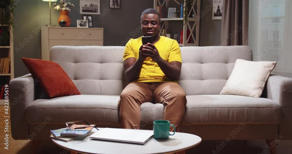 Single young Afro American relaxing, chatting with friends in living room at home. Attractive young guy sitting on couch and using smartphone in evening. Technology, social media concept. Single young Afro American relaxing, chatting with friends in living room at home. Attractive young guy sitting on couch and using smartphone in evening. Technology, social media concept.