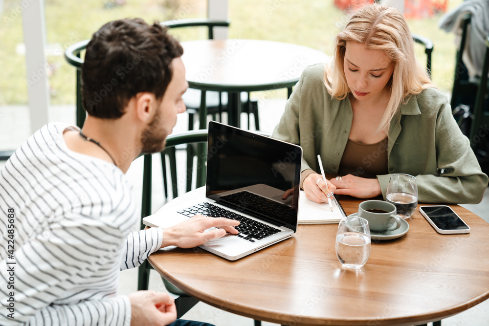 Man and woman using laptop and writing down notes while sitting in cafe Man and woman using laptop and writing down notes while sitting in cafe