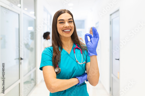 Young female medical doctor working at the hospital and medical staff  she is showing OK sign. Nurse stands in a walkway and smiles with her arms folded. She is wearing scrubs and a stethoscope.