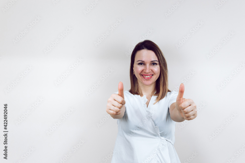 Beautiful young female doctor in a white medical gown, on an isolated white background. Cosmetologist smiles at the camera and holds thumbs up. With place for text or logo. Beautiful young female doctor in a white medical gown, on an isolated white background. Cosmetologist smiles at the camera and holds thumbs up. With place for text or logo.