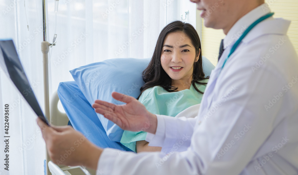 Obraz premium Young Asian medical man in eyeglasses showing chest X ray to smiling ethnic woman lying on sofa in hospital office.