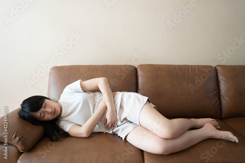 Young asian female suffering form stomachache while lying on couch at home. Causes of abdominal pain include menstruation pain, endometriosis, ovarian cyst, gastritis, colitis or IBS.