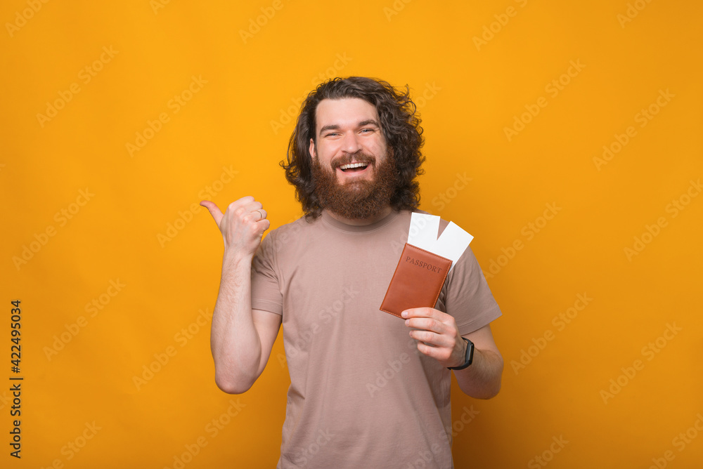 Photo of happy young man with beard holding passport with tickets and pointing away Photo of happy young man with beard holding passport with tickets and pointing away