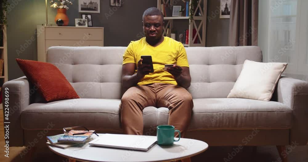 Attractive man sitting on sofa and entering card number on smartphone in living room. Afro American guy shopping online, buying clothes and making purchases in evening. Business concept. Attractive man sitting on sofa and entering card number on smartphone in living room. Afro American guy shopping online, buying clothes and making purchases in evening. Business concept.
