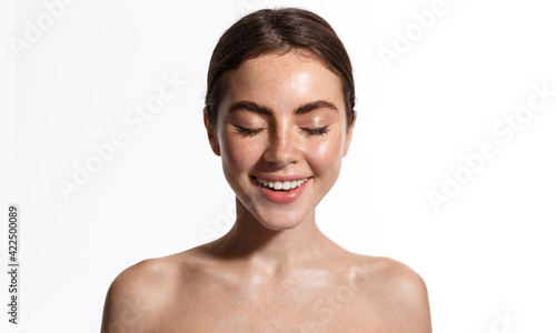 Beauty skin of young natural model with freckles, eyes closed. Girl trying new cosmetics, standing with naked shoulders, using facial toners and sunscreen spf nude makeup, white background