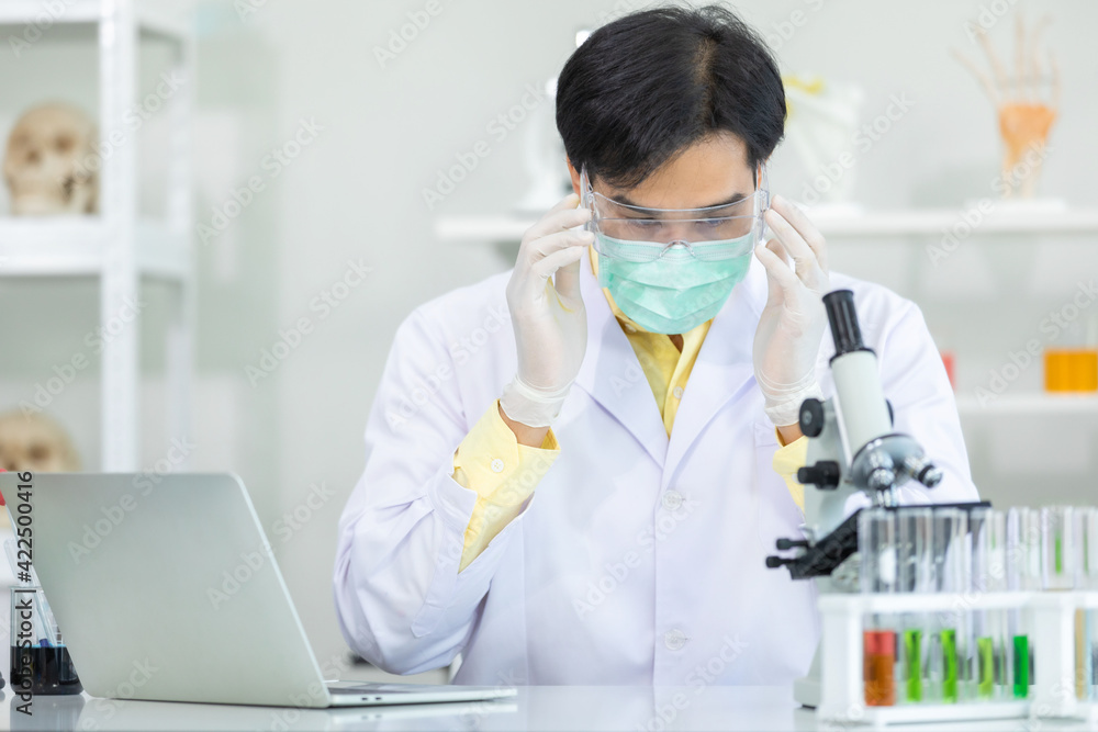 Asian man scientist wearing equipment mask and Protective glasses for working to computer laptop microscope while working on medical research in science laboratory. Concept research provent equipment. Asian man scientist wearing equipment mask and Protective glasses for working to computer laptop microscope while working on medical research in science laboratory. Concept research provent equipment.