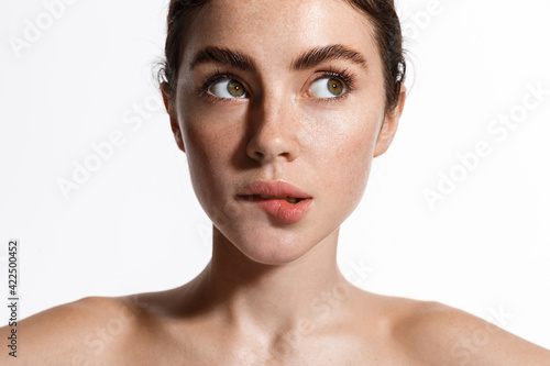 Beauty face and spa. Woman with freckles, clean nourished skin, biting lip and look aside. Girl model using antiaging cosmetics and vitamin c serum for bettet smoother skin tone, white background photo