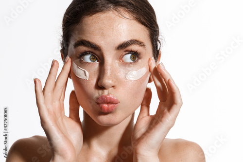 Close up beauty girl with freckles and thick eyebrows, applying moisturizing skincare cream, lotion or mask for skin lifting and anti-aging detoxifying effect, white background